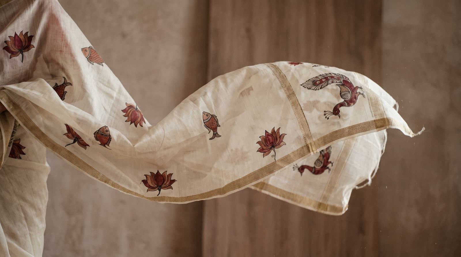 An ivory handloom saree hand-painted with fish, lotus and peacock motifs, draped and caught mid-air